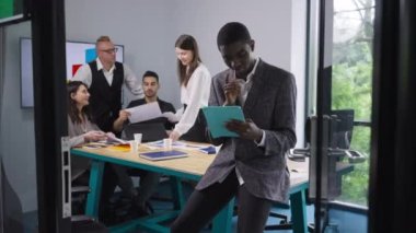 Düşünceli özümsenmiş Afrikalı Amerikalı erkek analist, arka planda beyin fırtınası yapan meslektaşlar olarak internette tablet üzerinde sörf yapıyor. Ofisteki e-ticaret başarısını analiz eden uzman bir adamın portresi.