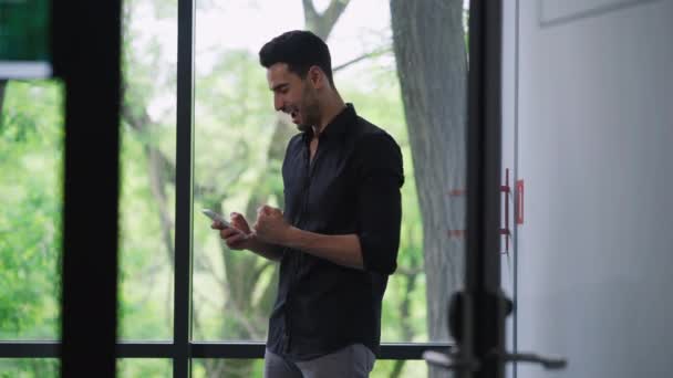 Emocionado hombre guapo de Oriente Medio haciendo gesto de victoria mirando a la pantalla del teléfono inteligente regocijando el éxito de inicio. Retrato del exitoso empresario confiado sonriendo mirando por la ventana en la oficina — Vídeos de Stock