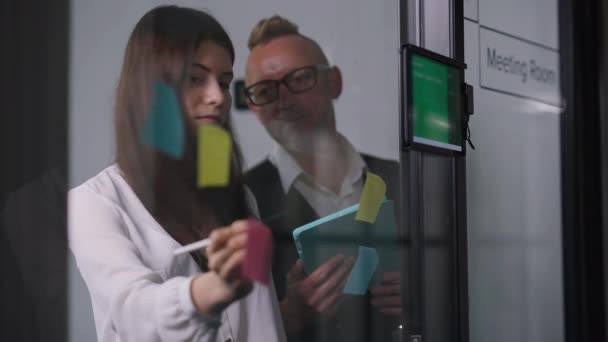 Mujer joven escribiendo con bolígrafo en notas adhesivas en la puerta de cristal como CEO masculina hablando de pie en la sala de reuniones. Mujer hermosa positiva y hombre guapo planificación de la estrategia de cooperación en la oficina. — Vídeos de Stock