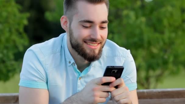 Lächelnder gutaussehender Mann schreibt Nachricht am Telefon im Park — Stockvideo