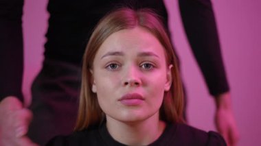 Pembe ışıklı stüdyoda elleri tarafından susturulmuş genç beyaz bir öğrenci, gözyaşı dolu bir ifade, yakın çekim, sessiz bir ağız, korku, hassasiyet, eller örtülü, dramatik ışıklandırma,