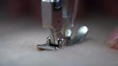 Kapalı makro görünüm, dikiş makinesinin ayaklarının yoğun desenli kumaş üzerinde süzüldüğünü gösteriyor. Parmaklar ise dikkatli kontrol ve hassas tekstil çalışmalarını vurgulayarak malzemeyi yönlendiriyor.