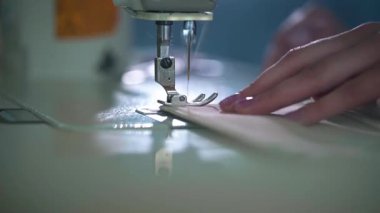 Bir el, dikiş makinesi iğnesinin altında yumuşak katlanmış kumaşı nazikçe yönetirken masadaki yansımalar ve sakin ışıklandırma dikkatli çalışmayı ve hassas tekstil işlemesini vurgular.