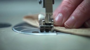 Yakın bir makro çekim, dikiş makinesinin ayağı kenar boyunca ilerlerken, katlanmış kumaşı sabit tutan parmakları gösterir, hassasiyet, doku ve dikkatli elle kontrol
