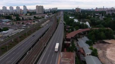 Geniş şehir otoyolu boyunca giden bir kamyon demiryolu rayları, konut blokları ve suyla yeşil alanların yanında, yukarıdan temiz bir şehir taşımacılığı manzarası yaratıyor.