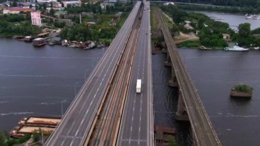 Bir yük kamyonu çok şeritli bir köprü boyunca ilerlerken, paralel bir demiryolu nehir boyunca ilerlerken, tekneleri, kıyı şeridi yapıları ve çeşitli kentsel manzaralar oluşturan yeşil alanlar da nehrin yanında yer alıyor.