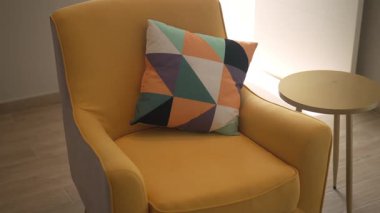 Renkli geometrik yastıklı sarı koltuk yumuşak doğal ışıkla aydınlık, modern bir evin içinde küçük yuvarlak bir masanın yanında duruyor..