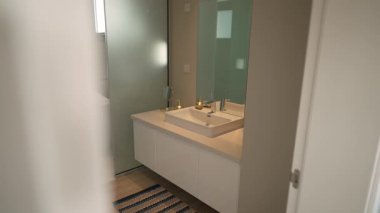 Modern banyo lavabosu aynası, yumuşak doğal ışık, serbest küvet görünür, fayanslı duş bölmesi, krom musluğu olan beyaz tezgah, ahşap zemin.