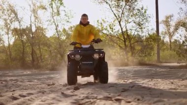 Solo ATV sürücüsü kum ve tozu tekmeliyor, sarı ceket, dönen lastik ve süspansiyonun yakın çekimi, güneş ışığı arka planı, dinamik arka plan hareketlenmesi