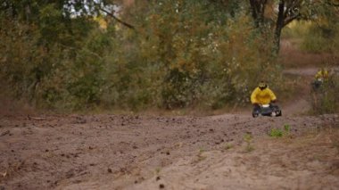 Ağır çekim. Sarı ceketli bir ATV binicisi çamurlu orman yolunda toz topluyor, uçan yapraklarla yüksek hızlı viraj, görünür koruyucu başlık ve eldivenler, sonbahar yaprakları