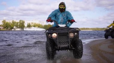 Ağır çekim. ATV rayları boyunca göl kumu ve çamur, sarı ceket ve kask, dar viraj kıvrımlı lastik izleri, su sıçramaları ve ıslak kıyı şeridi, bulutlu.
