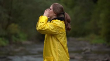 Sarı yağmurluklu beyaz kadın yardım istemek için ellerini kaldırıyor nemli orman zemininde, bulutlu ışıkta, yürüyüşçü rolünde, kararlı.