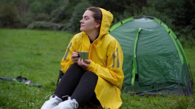 Sarı yağmurluklu kadın yeşil çadırın yanında çiğ çayırda kamp yapıyor, elinde fincanla çimenlere oturuyor, bulutlu gökyüzüne doğru bakıyor, yumuşak akşam ışığı, nemli dişliler.,