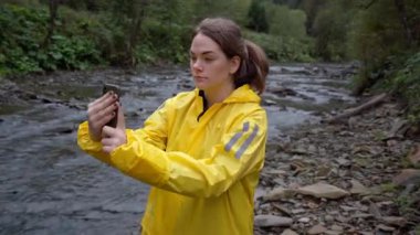 Sarı yağmurluklu beyaz kadın, Rocky River kıyısındaki telefonu kontrol ediyor, kamerayı selfie için ayarlıyor, yürüyüşçü ve bilim adamı gibi davranıyor, bulutlu ışık, ıslak kayalar.,