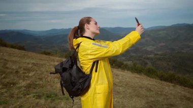 Sarı yağmurluk giymiş, dağ sırtında selfie çeken beyaz bir kadın. Sırt çantalı bir yürüyüşçü haritayı kontrol ediyor. Sonra panoramik video kaydediyor ve uzaktan el sallıyor.
