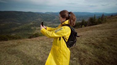 Sarı yağmurluklu bir kadın sırtta selfie panorama çekiyor, seyahat vlogger kayıtları dağ manzaralarını akıllı telefonla süpürüyor, sırt çantasıyla yürüyor, enerjik bir ruh hali içinde