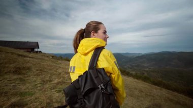 Sarı yağmurluklu kadın tepeden vadiye bakıyor. İzciler hava ve araziyi gözlerken gözleri, kırsal kabini ve kasvetli bulutları inceliyor.