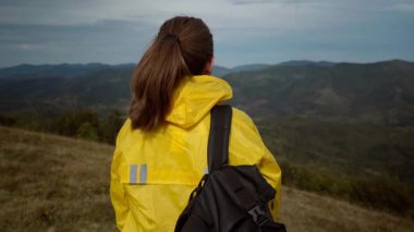 Alacakaranlıkta dağ manzarasına tepeden bakan sarı ceketli bir yürüyüşçü, çimenli bir sırt ve bulutlu gökyüzünün altında yuvarlanan tepeler, omzunun üstünden fırlatılan sırt çantası, yansıtıcı.