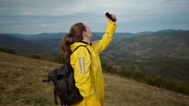 Sarı ceketli beyaz yürüyüşçü telefonu gökyüzüne kaldırıyor, panoramik dağ arkaplanı, siyah sırt çantası, rüzgarlı sırt, gülen yüz, geniş vadi,