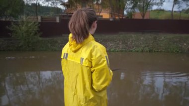 Nehir kıyısında duran sarı yağmurluk giyen bir kadının arka görüntüsü. Şiddetli yağmurdan sonra çamurlu, hızlı akan suya bakıyor.