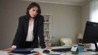 Ofis ortamında masasına yaslanan çekici genç bayan girişimci. Kameraya bakar, flört eder ve saçlarına dokunur.