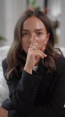 Siyah ceketli, ciddi bir genç kadının portresi doğrudan kameraya bakıyor. Elini yavaşça yüzünden çenesine kadar indirir.