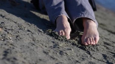 Çıplak ayaklı bir kadının kumdaki ayaklarına yakın çekim. Kumsalda oturuyor, ayak parmaklarıyla kumun dokusunu hissediyor ve dinleniyor.