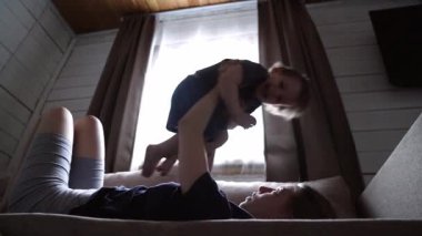 Anne göğsünün üstünden bebek kaldırıyor, arka ışıklandırmalı pencereler, yumuşak perdeler çerçeveli ahşap yatak odası, ebeveyn yatağa uzanıyor gülümseyen çocuğu destekliyor, kıkırdama hareketi, şakacı kucaklaşma ve göz teması.,