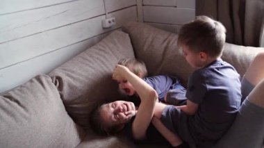 Kafkas anne ve iki çocuk kanepede gıdıklanıyor, güneşli ahşap oturma odası, içten bir kahkaha, nazik güreş, kardeş yaramazlığı, ebeveyn sabrı, rahat yastıklar, sıkı sarılma, şakacı kaos, ev