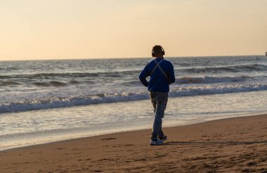 Gün batımında ya da gün doğumunda müzik dinleyerek sahilde mutlu yürüyen bir insan.