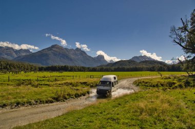 Campervan crossing an unbridged stream. Glenorchy (near Queenstown), South Island, New Zealand. Lush green meadows in a wide valley, with impressive mountains in the background. clipart