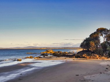 Waterhouse Koruma Alanı, Bridport, Kuzeydoğu Tazmanya, Avustralya 'daki ıssız sahilde akşam güneşi. Kayalık çıkıntılar, uzaktaki tepeler