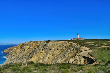 Cape Espichel Deniz Feneri (Farol do Cabo Espichel), turkuaz Atlantik Okyanusu 'ndaki dramatik kıyı uçurumlarının tepesinde berrak mavi gökyüzünün altında, Sesimbra, Portekiz.
