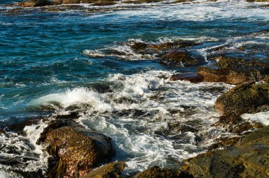 Okyanus dalgaları, parlak güneş ışığı altında engebeli, ıslak kıyı kayalarına çarpıyor. Güney Avustralya, Fleurieu Yarımadası.