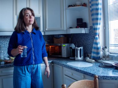 Parlak mutfağında elinde bir bardak suyla mavi aksanlı bir kadın duruyor. Sabah ışığı pencereden süzülüp güne hazırlanırken huzurlu bir atmosfer yaratıyor..