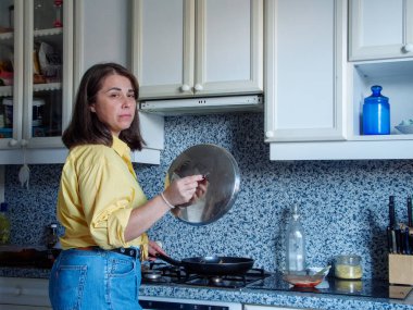 Bir kadın sıcak bir mutfakta elinde bir tavayla dikiliyor. Güneş ışığı boşluğu dolduruyor, lezzetli bir yemek hazırlarken odaklanmış yüz ifadesini vurguluyor..