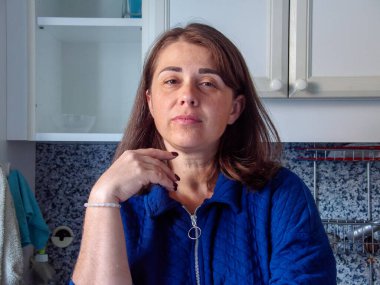 Kahverengi saçlı ve mavi ceketli bir kadın parlak bir mutfakta oturur ve yemeği hazırlarken dikkatlice uzaktan bakar. Sıcak öğleden sonra güneşi odayı yumuşak ışıkla dolduruyor..