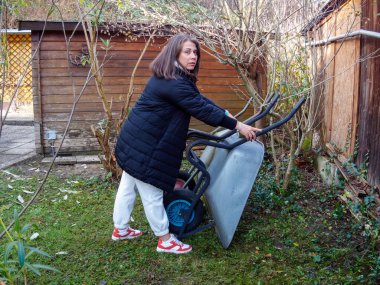 Uzun saçlı bir kadın yemyeşil bir bahçede el arabasını itiyor. Güneş yavaşça parlıyor ve yakınlardaki yeşil ve ahşap yapılarda dolaşırken huzurlu bir atmosfer yaratıyor..