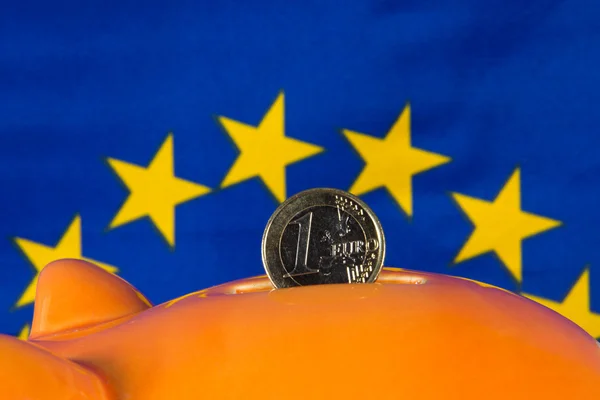 Prasátko s jedno euro mince, vlajka Eu v pozadí — Stock fotografie