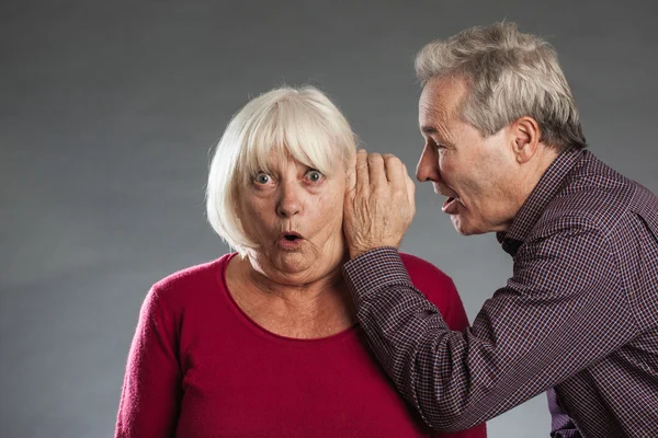 Seniorenpaar, Mann flüstert Frau ins Ohr, sie sieht überrascht aus. — Stockfoto