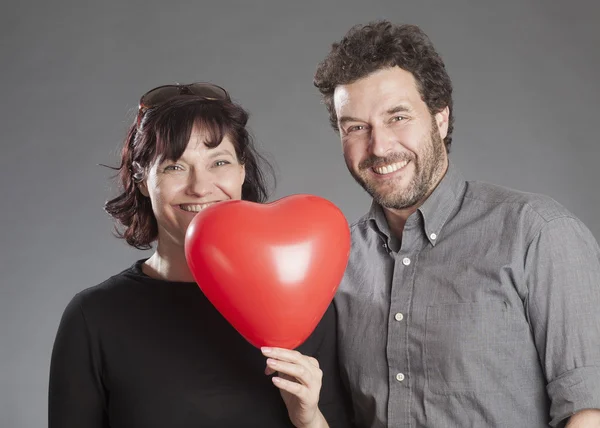 Pareja madura sonriendo sosteniendo el globo rojo en forma de corazón — Foto de Stock