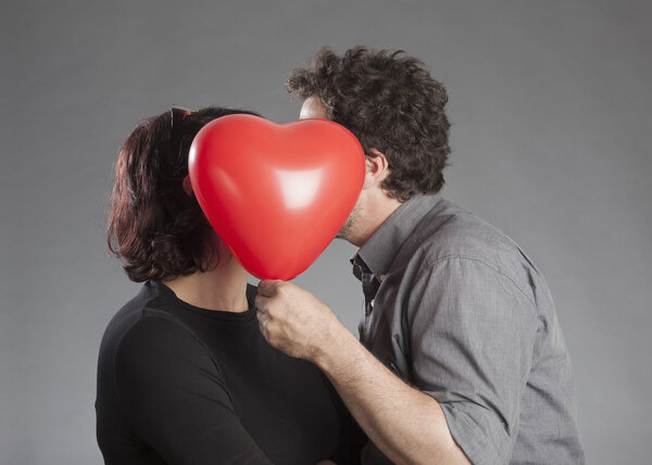 Зрелая пара улыбается, прячась за красным шаром в форме сердца.
