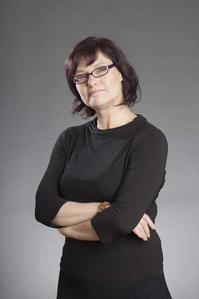Reife brünette Frau mit Brille steht mit gefalteten Händen vor grauem Hintergrund — Stockfoto