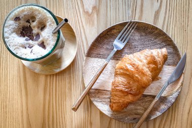 Kahvaltı takımı, kruvasan ekmeği, kahverengi ahşap tabakta buzlu kahve ve masa.