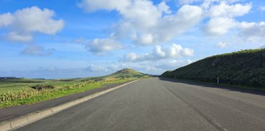 Yuvarlanan tepeler ve kabarık bulutlarla dolu canlı bir gökyüzü tarafından çerçevelenmiş açık bir yol ufka doğru uzanır.