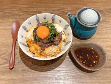 Lezzetli Gyudon, çiğ yumurta sarısı ve dilimlenmiş pirinçli biftek, yanında sos ile servis edilir.