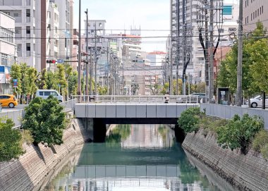 Beton duvarlar ve yemyeşil yemyeşil ile çevrili, yüzeyi açık bir gökyüzünün altında yansıtılan binalar ve elektrik direkleri ile huzurlu bir kanal bir Japon şehrinin içinden akar.