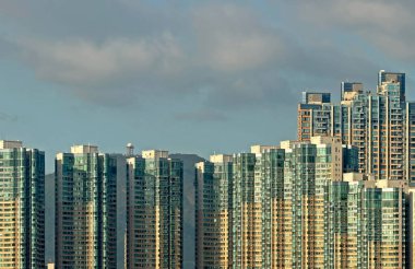 Tepelerin ve parçalı bulutlu gökyüzünün zemininde yansıtıcı cam pencereler ve balkonlara sahip çok sayıda çağdaş yerleşim kulelerinin yer aldığı yoğun bir kentsel manzara.