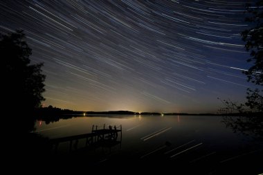 Yıldız izleri ve uydu izleri gece gökyüzüne doğru kendi çizgilerini çiziyor. Aynı zamanda sakin sulara da yansıyor..