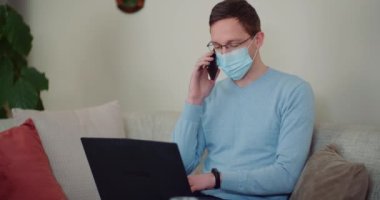 Dizüstü bilgisayarda çalışırken koruyucu maske takan ve cep telefonuyla konuşan adam.